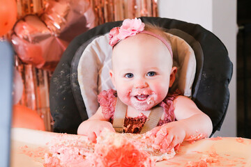 Cute birthday girl celebrating her one year birthday by messily eating a cake surrounded by pink background streamers and balloons