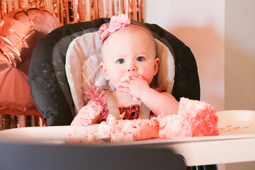 Cute birthday girl celebrating her one year birthday by messily eating a cake surrounded by pink background streamers and balloons