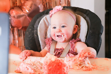 Cute birthday girl celebrating her one year birthday by messily eating a cake surrounded by pink background streamers and balloons