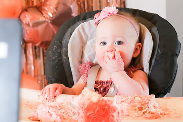 Cute birthday girl celebrating her one year birthday by messily eating a cake surrounded by pink background streamers and balloons