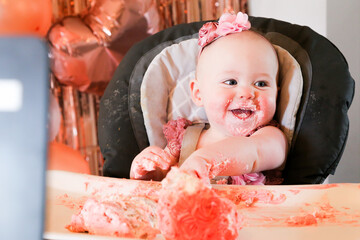 Cute birthday girl celebrating her one year birthday by messily eating a cake surrounded by pink background streamers and balloons
