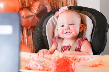 Cute birthday girl celebrating her one year birthday by messily eating a cake surrounded by pink background streamers and balloons