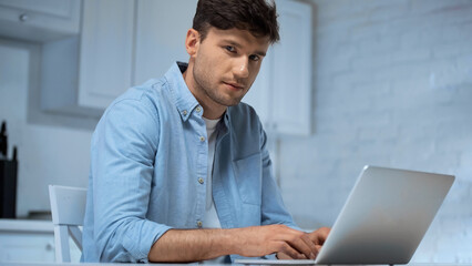 freelancer in blue shirt looking at camera while using laptop in kitchen
