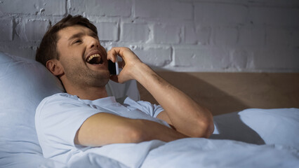 excited man laughing while talking on mobile phone in bed