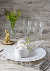 A white egg decorated with rabbit ears lies on a white plate. A vase of flowers, two glass glasses and a rabbit-shaped candle in the background. Easter Lunch