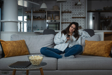 Obraz premium Young pretty woman sitting at home on the lazy weekend watching television feeling bored. Female in cozy apartment enjoying free time while her boyfriend or husband working.