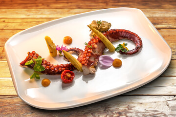 Roasted octopus tentacles with young baby corn, tomato and mint leaves in white plate on wooden boards
