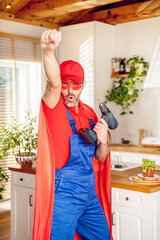 Home hero. A husband dressed as a superhero fixes furniture in the kitchen and poses with a drill...