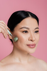 Asian woman with naked shoulders massaging cheek with jade roller isolated on pink