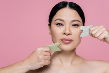 Asian woman scraping face with jade stones and looking at camera isolated on pink