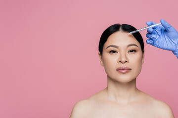 Cosmetologist in latex glove holding syringe near forehead of asian woman isolated on pink