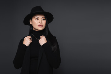 Fashionable asian woman in fedora hat looking away isolated on dark grey