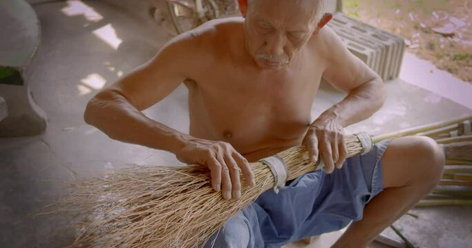 Asian male farmer who is an elderly person makes brooms from the grass that has many names called Paddy's lucerne, Queensland hemp , Arrowleaf sida , Common sida , Cuba juite for extra income.
