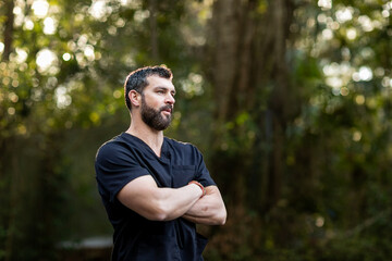 A doctor with dark hair and a beard in black scrubs standing outside in a natural green environment