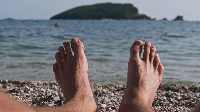 POV view of male barefoot feet on the beach by the sea. Legs of a young man lying on a pebble beach by the calm water. Funny male legs sunbathe and relax on the tropical resort. Summer exotic vacation