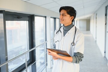 Portrait of confident young Arabian Indian male medical doctor in white coat, standing with clipboard in hands on background of modern hospital building outdoor