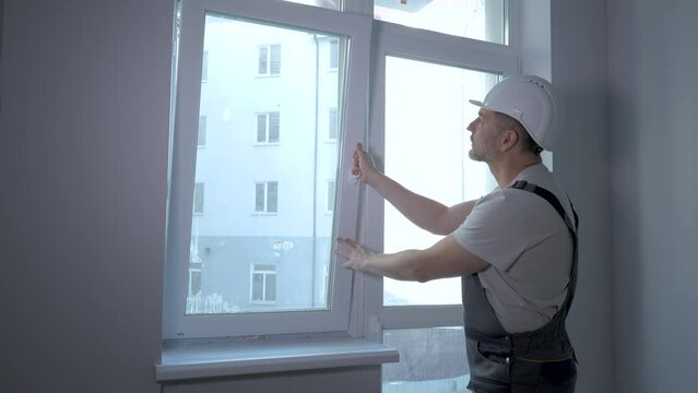 A builder in uniform checks the size and quality of window installation in a new apartment