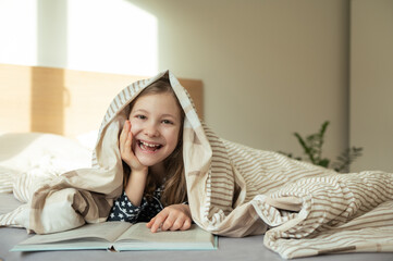 Cute teen child girl lying and reading book on bed under the blanket