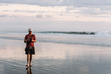 Latino male photographer walking on the beach looking at his camera, wearing a cap on his head.