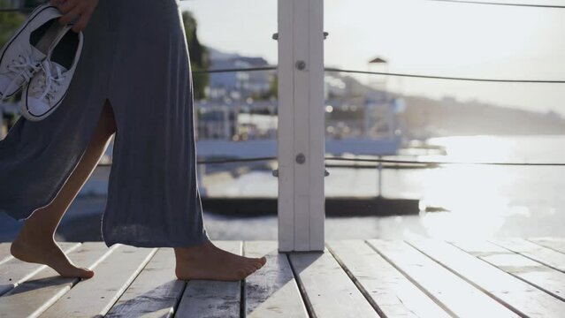 Unrecognizable barefoot young woman walks along wooden pier holding sneakers in hand enjoying sunny morning on seaside, detail view female feet against beach at blurred background