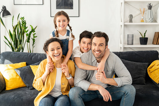 Happy together. Portrait of cheerful multiracial family of four - mom, dad, son and toddler daughter sitting on the sofa in cozy living room, looking at camera and laughing, enjoying weekend at home