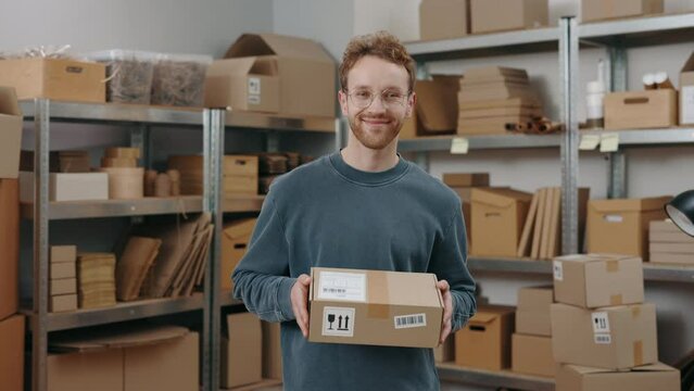 Small business. Waist up portrait view of the stylish guy wearing glasses standing at the warehouse and looking at the camera while holding parcel. Occupation concept.