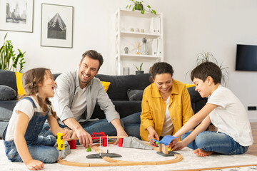 Lovely family of four spend weekend together at home, parents and two kids playing toys, sibling building railway, have a fun. Mom, dad and children sitting on floor in cozy living room and playing