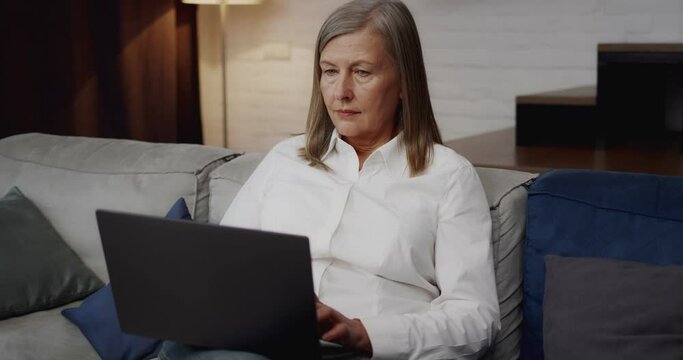 Attractive Mature Businesswoman Texting Message on Modern Laptop sitting at Home. Middle Aged 60s Lady Working Remotely, Chatting looking at notebook computer screen. Senior Progressive Person.