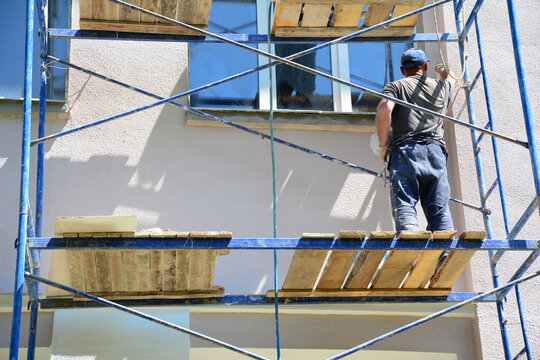 Painting of a stucco exterior wall of a house. A building contractor on scaffolding is painting, renovating a stucco facade.