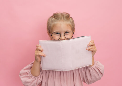 Little girl in eyeglasses holding book album or diary on pink background.