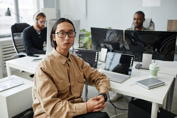 Young serious Asian businessman in eyeglasses and casual clothes sitting by desk with two laptop...