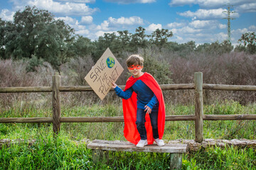 Little Boy Pretending To Be A Superhero With A Red Cape, Holding A Cardboard Sign That Says SAVE...