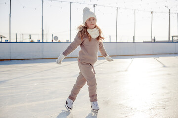 Funny little blonde girl of 7 years old in casual clothes posing on a skating rink in skates. The...