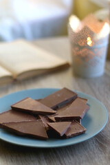 Plate of chocolate, open book, reading glasses, lit candle and flowers on the table. Selective focus.