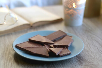 Plate of chocolate, open book, reading glasses, lit candle and flowers on the table. Selective focus.