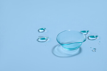 One contact lens and drops of water on light blue background. Space for text