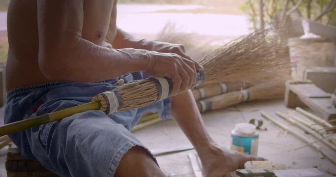 Asian male farmer who is an elderly person makes brooms from the grass that has many names called Paddy's lucerne, Queensland hemp , Arrowleaf sida , Common sida , Cuba juite for extra income.