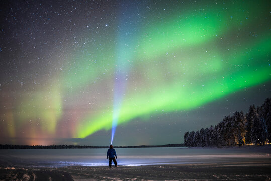 Person under Northern Lights display (aurora borealis) having amazing travel experience with amazing bright colourful green and purple night sky and stars in Lapland, inside Arctic Circle in Finland