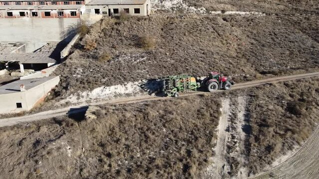 Tractor rojo circulando por un camino en la monta&ntilde;a, transportando una m&aacute;quina para esparcir mineral en los campos. Video a&eacute;reo