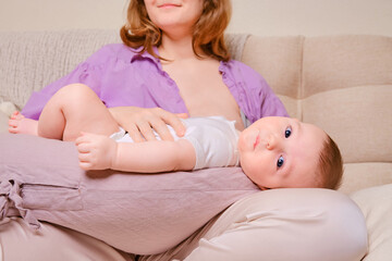 Woman mother breastfeeds an infant baby sitting on a home sofa. Mom feeds milk toddler baby in...
