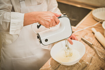 Adult woman in white chef clothes beats eggs with mixer for making pie in home kitchen