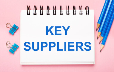 On a light pink background, light blue pencils, paper clips and a white notebook with the text KEY SUPPLIERS