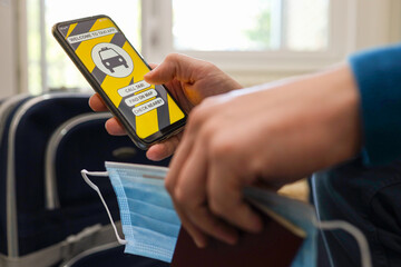 Young man ready to travel, calling a taxi via taxi app. Holding passport and covid mask with suitcase in background. Businessman, traveler, student concept.	