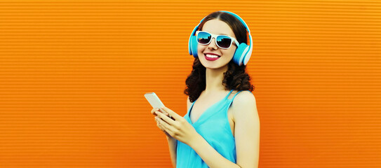 Portrait of happy smiling young woman in headphones listening to music with smartphone on orange background