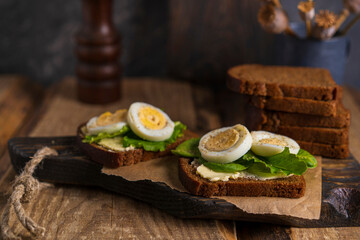 Black whole grain sliced bread with butter, salad leaves  and boiled eggs on wooden rustic table. Country, organic dietic opened sandwich. Copy space. Dark backgroound.