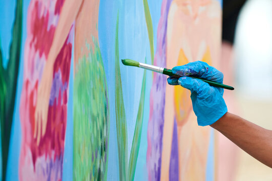 Artist's hand in blue gloves with paintbrush painting colorful picture at outdoor art festival