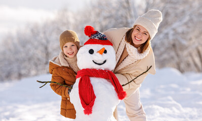 Joyful young mother making snowman with son while playing together in snowy winter park