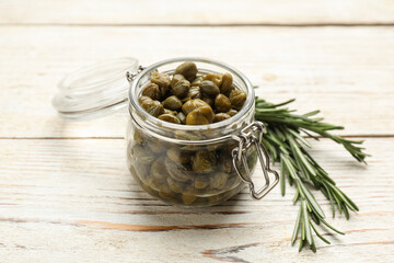 Tasty capers in glass jar and rosemary on white wooden table