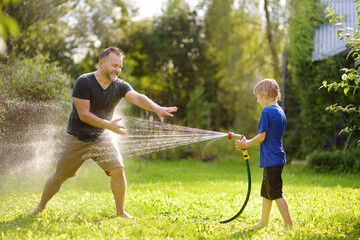 Funny little boy with his father playing with garden hose in sunny backyard. Preschooler child having fun with spray of water. Summer outdoors activity for kids. - obrazy, fototapety, plakaty