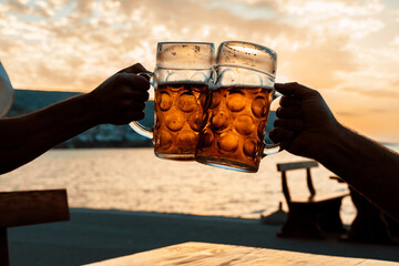 Cold beer in the hands, toast with a mug of beer on the coast in the sunset. Summer, Party, Holiday, 